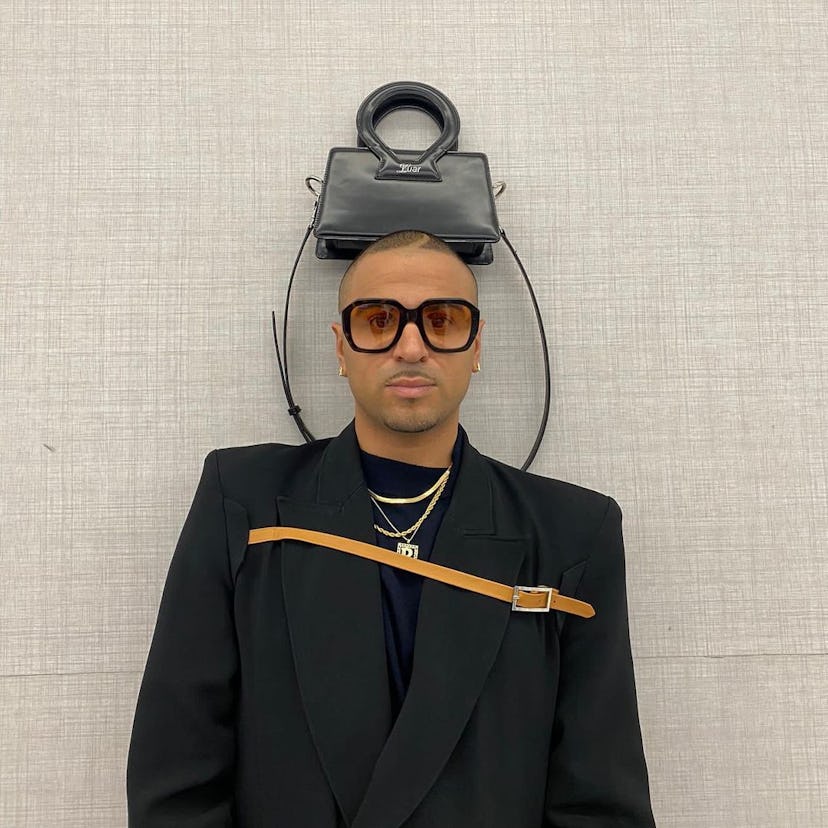 Man wearing glasses, a black blazer with a brown belt, and a black Luar bag on top of his head