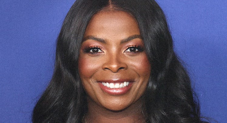 A smiling woman with long wavy hair, wearing a mustard yellow top and a necklace, against a blue bac...