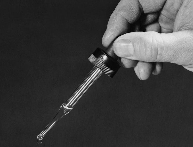 A black and white image of a hand holding a pipette with a drop of Facial Oil dripping.