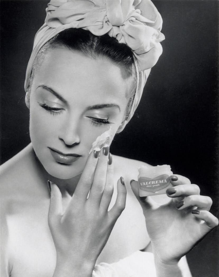 Woman applying skin cream, 1945-1955. Glamour fashion shot of a model applying Valcrema skin youth t...